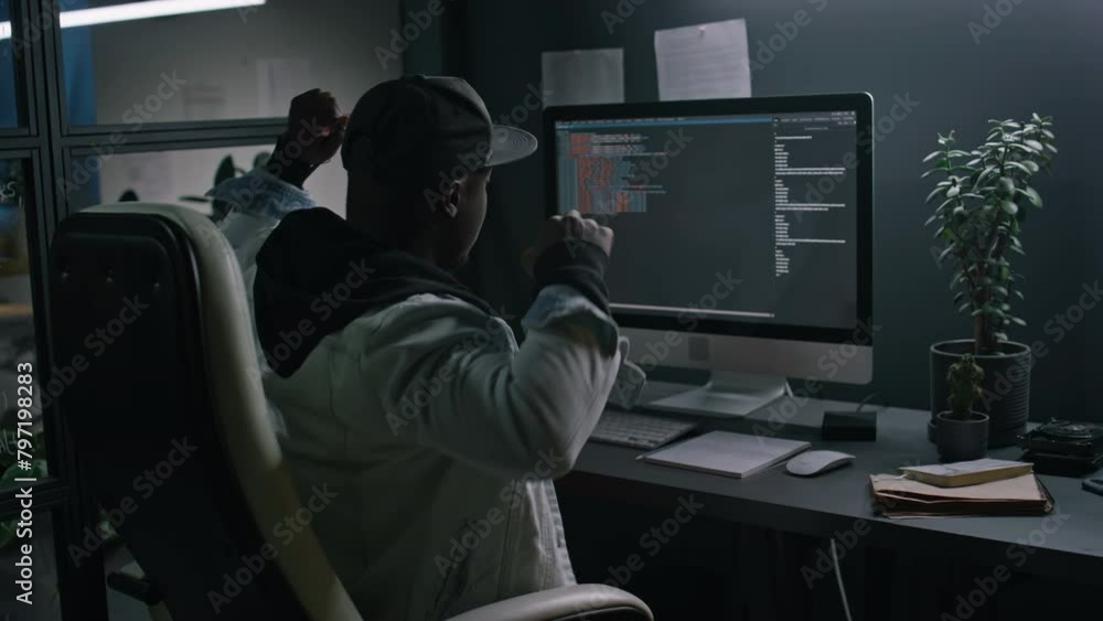 Medium rear of tired young African American male programmer in hoodie, baseball cap working on computer code in dark office late at night, stretching and rubbing eyes Medium rear of tired young African American male programmer in hoodie, baseball cap working on computer code in dark office late at night, stretching and rubbing eyes
