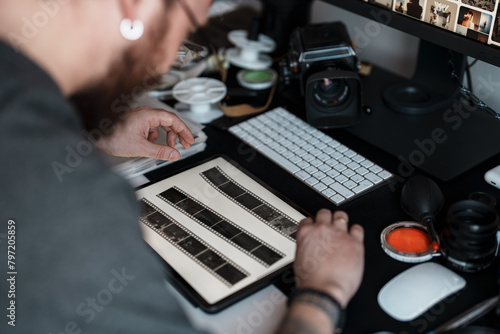 Focused photographer examines film strips amidst a modern workspace setup with a digital workstation and vintage camera equipment. photo