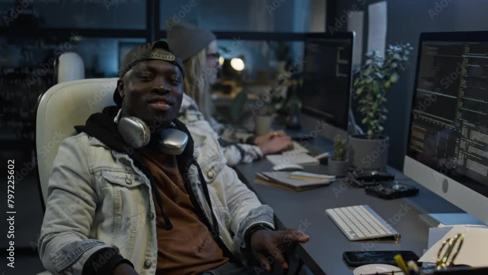 Medium portrait of cheerful young African American man sitting at desk in IT startup office, with team colleagues, writing computer code, turning towards camera and smiling Medium portrait of cheerful young African American man sitting at desk in IT startup office, with team colleagues, writing computer code, turning towards camera and smiling