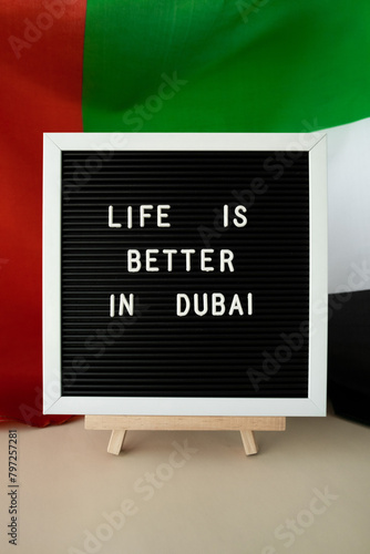 Message LIFE IS BETTER IN DUBAI on background of waving UAE flag made from silk. United Arab Emirates flag with concept of tourism and traveling. Inviting greeting card, advertisement. Dubai welcoming