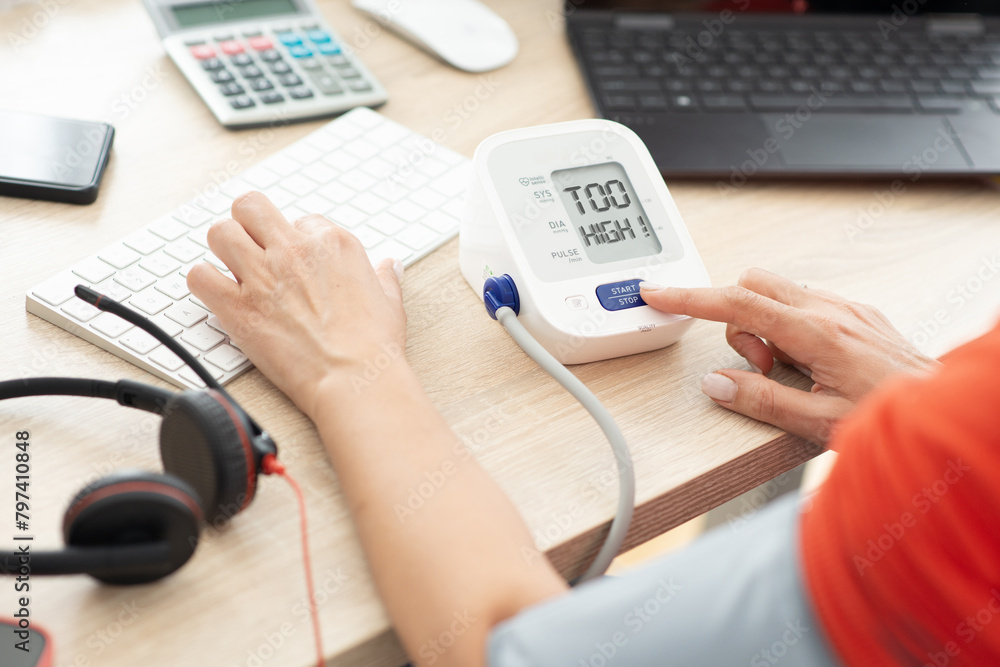 Hypertension due to office stress concept with woman working at computer desk taking blood pressure with a medical device Hypertension due to office stress concept with woman working at computer desk taking blood pressure with a medical device