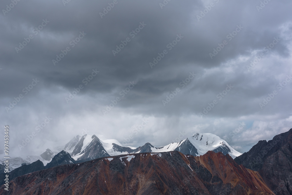Dark atmospheric alpine landscape with sharp rocky ridge and snowy peaked tops in low heavy lead gray cloudy sky. Rock mountain wall and snow-capped pointy peaks in bleak dramatic bad rainy weather.