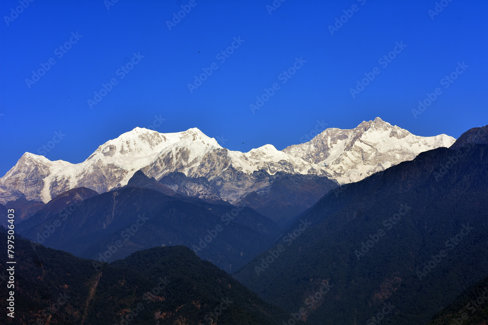 Majestic snow-capped mountain range under a clear blue sky, with forested foothills in the foreground. Mt. Kangchenjunga and its ranges from Sikkim