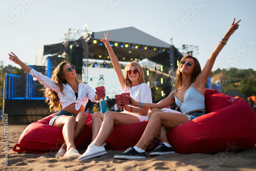 Girls relax on red bean bag chairs at beach music festival. Group enjoys cold drinks, summer vibes, ocean backdrop. Carefree atmosphere, casual fashion, youth enjoying live entertainment, sea view. © artiemedvedev