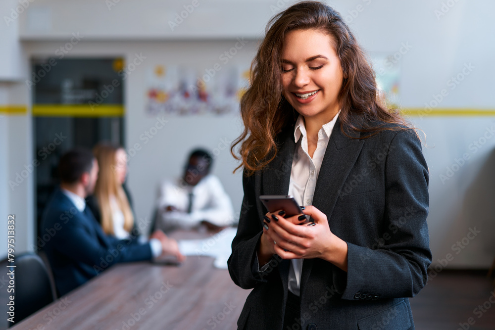 Businesswoman in smart casual attire smiles at smartphone in office setting team discussion blurred in background. Pro multitasking connects online stays informed with tech corporate environment. Stoc...