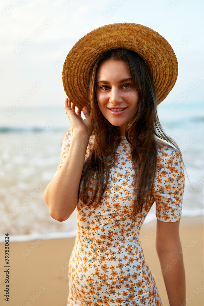 Smiling woman in floral dress and straw hat on sunny beach, gentle sea waves behind. Fashionable summer attire, retro vibes, travel leisure concept. Happy female enjoys coastal ambiance, warm light.
