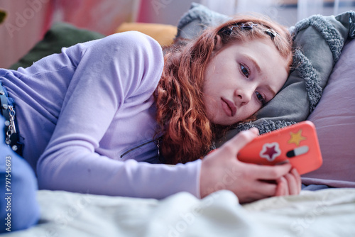 Melancholic teen girl lying on bed staring at smartphone screen waiting for message or phone call