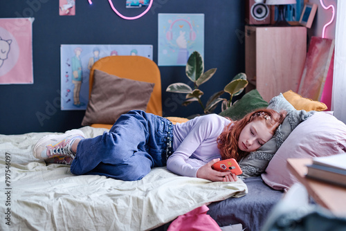 Modern Caucasian teen girl spending day indoors relaxing on bed in her bedroom watching videos in social media on smartphone
