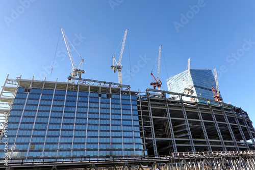 Several tower cranes are building a modern tall skyscraper against a clear blue cloudless sky