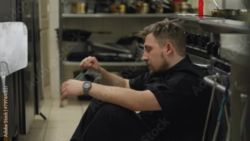 Close-up of a tired male cook in a black uniform takes off his hat and straightens his hair while sitting on the floor leaning on the table in the kitchen of a restaurant photo