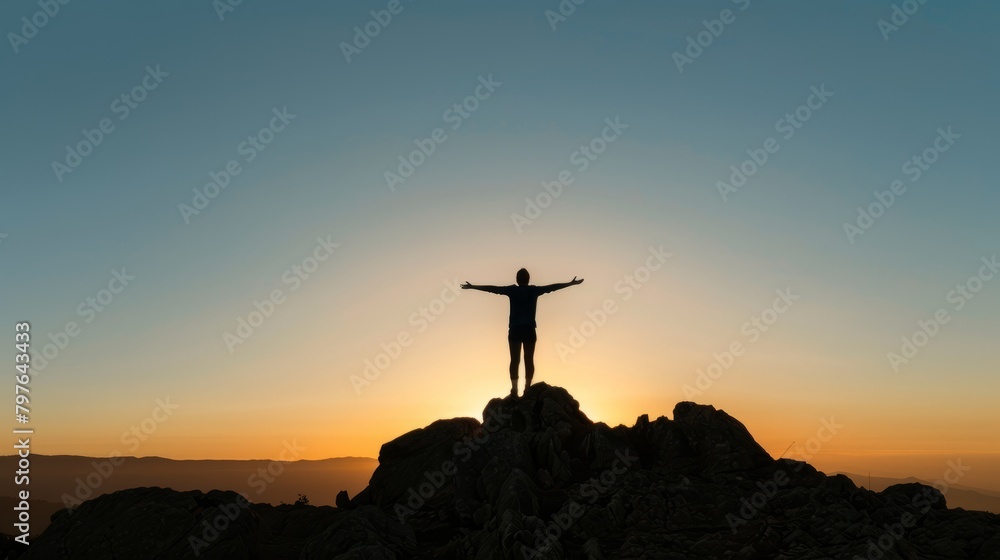 A silhouette shot at sunset of a young person standing on a mountaintop with outstretched arms, enjoying the view from the top