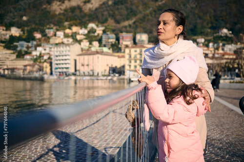 Young pretty woman and her cute daughter , a mother and little child girl in warm winter clothes, standing together on the promenade, admiring the beauty of the lake of Como in Italy