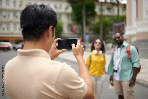 Smartphone in hands of young male tourist guide taking picture of intercultural couple standing in urban environment