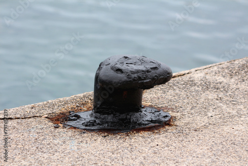 Vintage old restored and freshly painted black old iron mooring bollard screwed on local concrete pier with four strong metal bolts surrounded with rust dust left from cleaning before painting