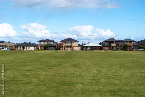 Background texture of a large vacant public sports ground with green grass lawn with some modern residential suburban houses in the distance. An outdoor park in Point Cook, Melbourne VIC Australia. © Doublelee