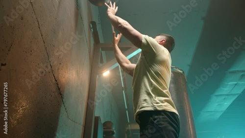A man in a focused squat position throws a medicine ball against a wall, demonstrating strength and endurance in a gritty gym setting during an intense cross-training session. Camera 8K RAW. 