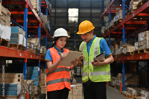 Two warehouse workers wearing safety hats are holding board and checking shipping stocks in storage, warehouse, or factory for delivery, Commercial Industry Business Concept.