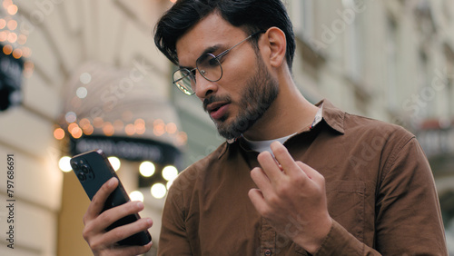 Close up upset unhappy Indian Arabian ethnic man young guy businessman male using looking mobile phone bad connection losing fortune loss fail touch head why negative result outdoors city urban street