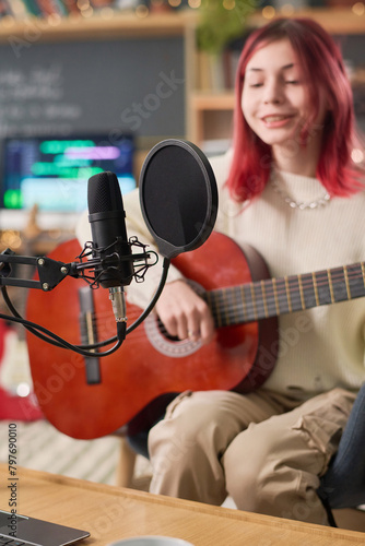 Focus on microphone fixed on desk in front of camera against talented teenage girl playing acoustic guitar and singing song while recording it