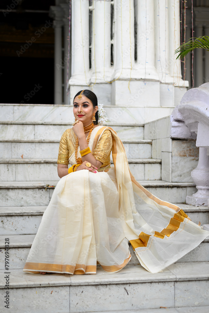 Stunning bride dressed in a traditional Indian bridal saree with gold jewellery smiles tenderly sitting on the staircase of a vintage building. Wedding fashion and lifestyle.