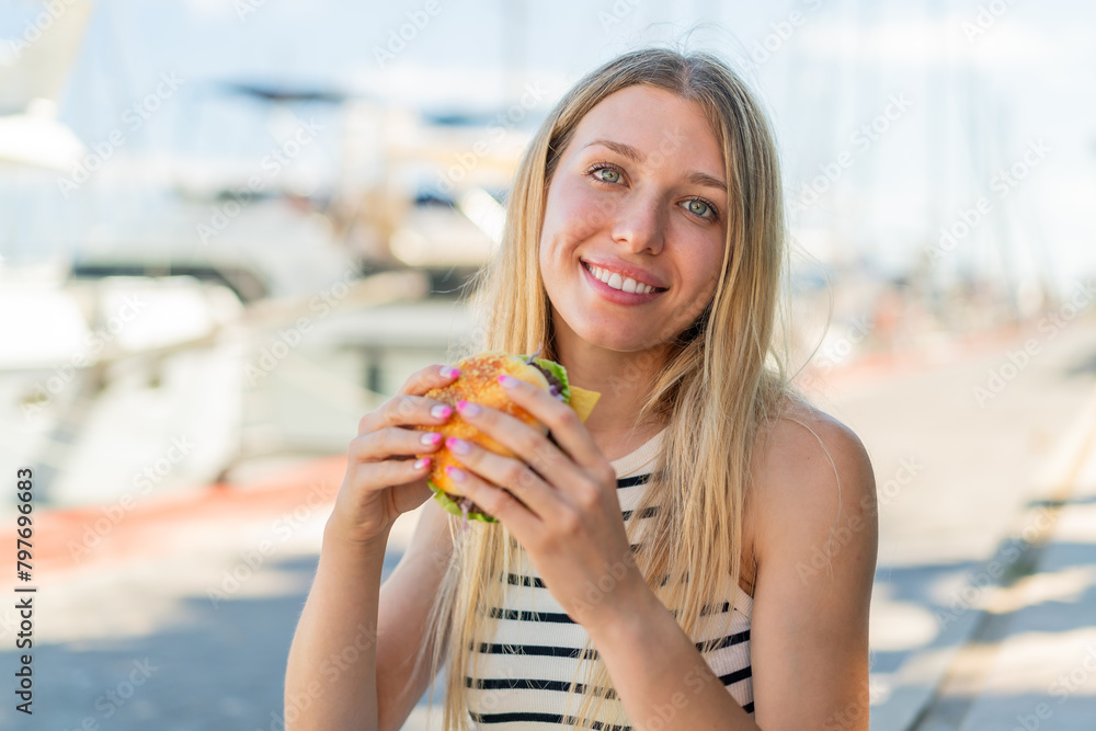 Young blonde woman at outdoors holding a burger with happy expression Young blonde woman at outdoors holding a burger with happy expression