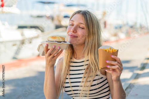 Young blonde woman at outdoors holding a burger and fried chips with happy expression
