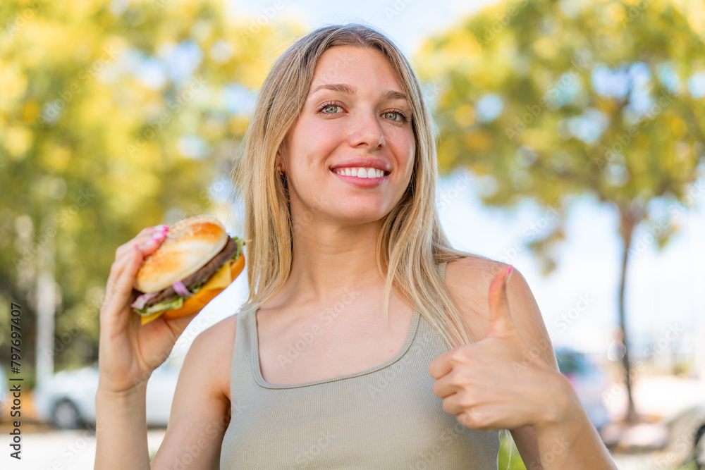 Young blonde woman holding a burger at outdoors with thumbs up because something good has happened Young blonde woman holding a burger at outdoors with thumbs up because something good has happened