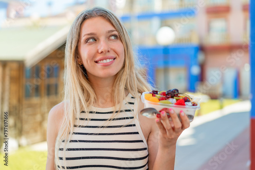 Young blonde woman holding a bowl of fruit at outdoors looking up while smiling