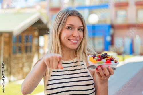 Young blonde woman holding a bowl of fruit at outdoors points finger at you with a confident expression