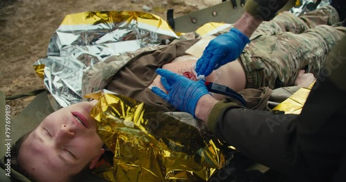 A confident male medic in blue medical gloves stops the blood of a wounded soldier in a camouflage uniform who is lying on a military medical stretcher in the combat area photo