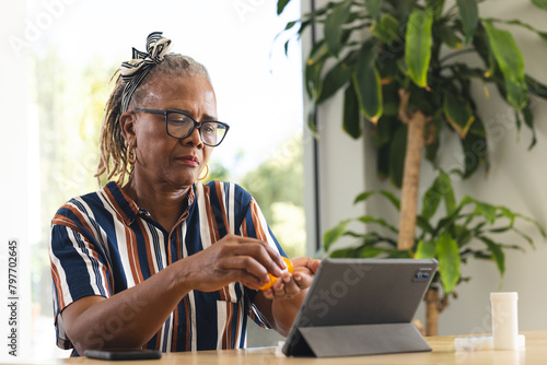 A senior African American woman wearing glasses is using a tablet at home and taking medicine, copy  photo