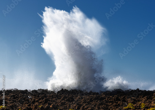 wave crashing onto a rocky reef under a blue sky and creating an eruption of water photo