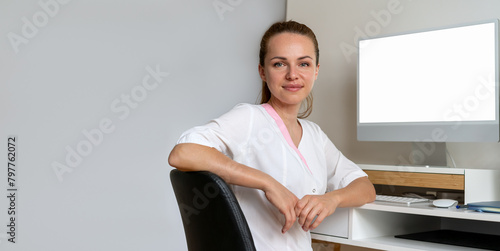 Portrait of a brunette medical worker in a white lab coat sitting at a desk with a computer and performing medical administrative tasks.