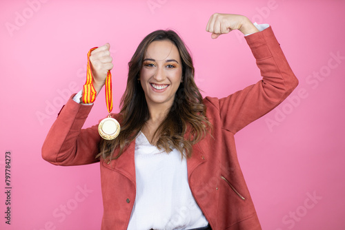 Young beautiful woman wearing casual jacket over isolated pink background holding a medal showing arms muscles smiling proud