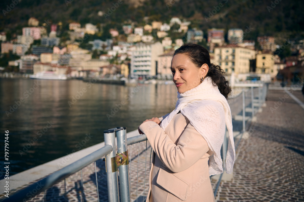 Portrait of a beautiful young woman in elegant beige coat, smiling and dreamily looking away, walking on the promenade of The Como lake over Alpine mountains background. Wonderful travel destination