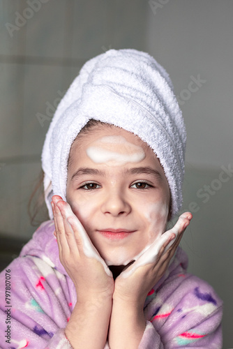 A girl in the bathroom with a towel on her head washes her face with cleansing foam with her hands and smiles