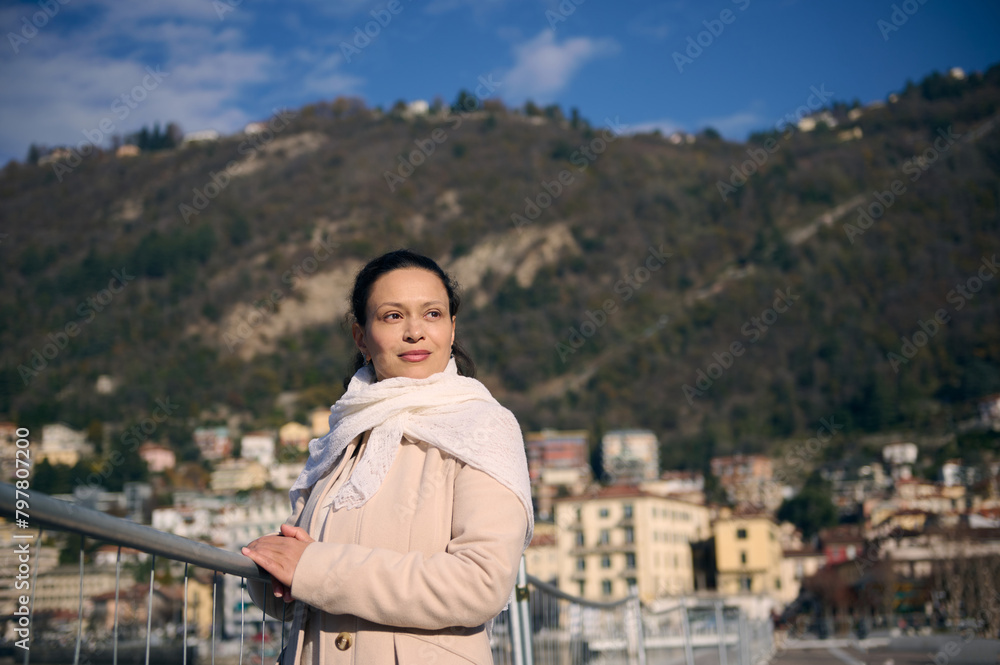 Multi ethnic young woman smiling on the lakefront, dreamily looking into the distance. Portrait of a beautiful brunette in beige coat, against the Alpine mountains on background in the city of Como