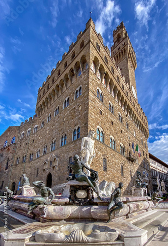 Florence, Italy - April 05, 2023 : view of Palazzo Vecchio with statue of Neptune with other mythical figures on octagonal fountain (16th century) in Piazza della Signoria on beautiful blue sky