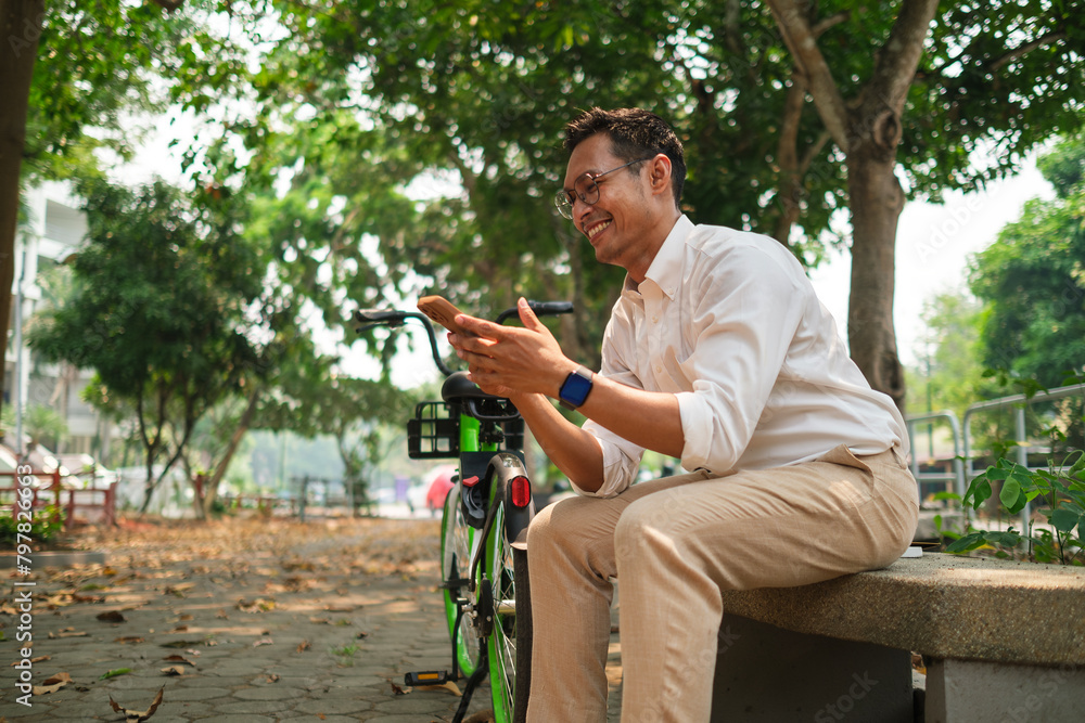 Happy adult businessman sitting on a bench next to his bicycle in city park and using his mobile phone. Happy adult businessman sitting on a bench next to his bicycle in city park and using his mobile phone.