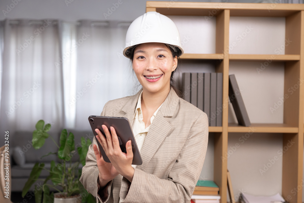 Asian project manager or architect female wearing hardhat holding tablet talking to customers or assigning work to employees, start-up contractor or engineer worker woman joyful working at home office