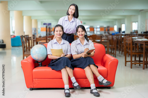 Full body image of a group of 3 Asian female students sitting on a red sofa. Holding a textbook and a tablet with a globe model next to a geography subject It is in the library of a famous high school