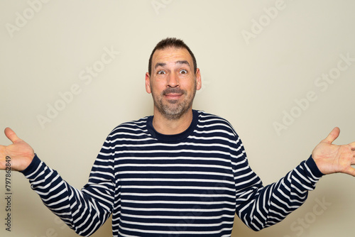 Hispanic man with beard in his 40s wearing a striped sweater clueless and confused with open arms, no idea concept. Isolated on beige studio background.