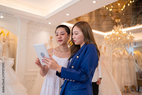 Asian bride is smiling while choosing wedding dress in modern wedding salon, Attractive designer girl using tape meter fitting on wedding dress at wedding studio.