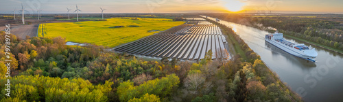 Ground-mounted solar photovoltaic panels, wind turbine farm neighboring canal, where ship gracefully navigates waterway. Photovoltaic park, windmill park, rapeseed field   freight ship on Kiel Canal. photo