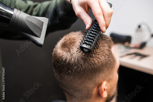 Barber brushes man short hair using dryer in barbershop. Skilled barber does elegant hairstyle in beauty salon. Male haircut