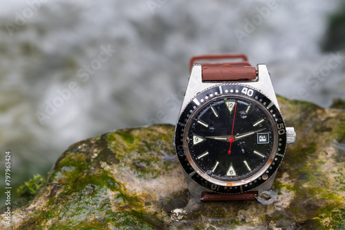 A used, sturdy outdoor watch lies on a rock by the rushing stream, embodying unwavering reliability for every outdoor adventure.