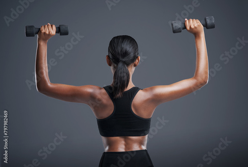 Back, fitness and woman with dumbbell in studio for muscle workout, training and healthy body. Sports, person and athlete with equipment for cardio, strength exercise and wellness on gray background