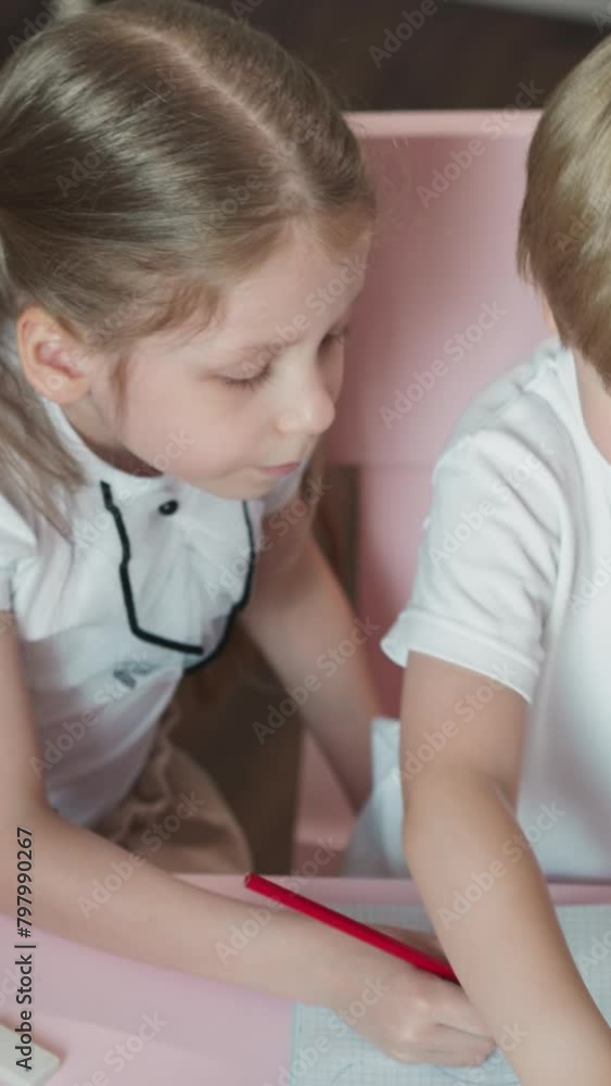 Caring sister draws picture for little brother sitting at desk at home. Little girl shows exercise to junior boy playing school together in children room Caring sister draws picture for little brother sitting at desk at home. Little girl shows exercise to junior boy playing school together in children room