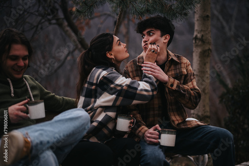 An affectionate couple is playfully interacting with each other  while a friend laughs beside them  all sharing a moment in a cozy outdoor setting.
