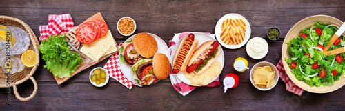Summer BBQ or picnic table scene with hamburgers, hotdogs, salad and snacks. Top down view over a dark wood banner background.
